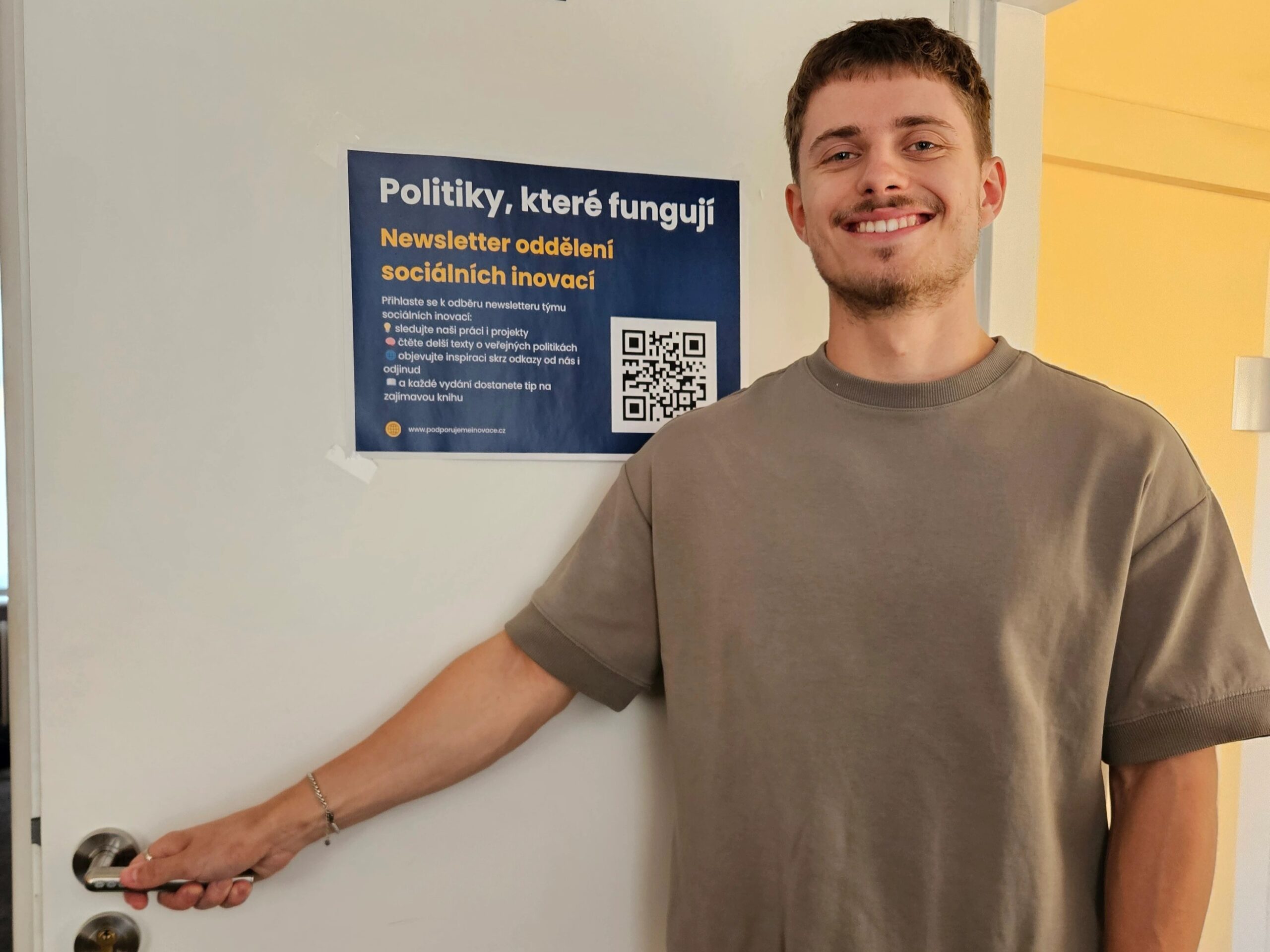Mladý muž a stážista autor tohoto textu, stojí v šedém tričku u bílých dveří, drží kliku a usmívá se do kamery. Na dveřích je vylepený plakát s nápisem „Politiky, které fungují“ a podtitulem „Newsletter oddělení sociálních inovací“. Na plakátu je také QR kód a odkaz na webovou stránku.