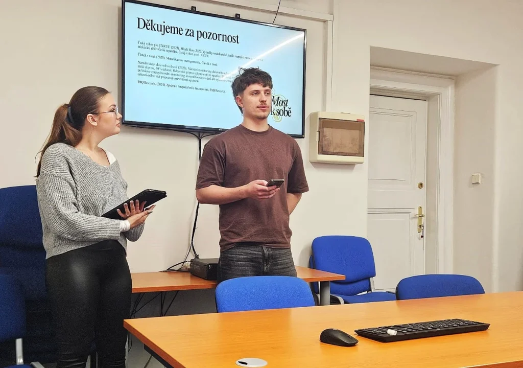 Student Honza Veselý v hnědém tričku stojí uprostřed zasedací místnosti a v ruce drží smartphone. Po jeho levici stojí kolegyně v šedém svetru s tabletem a oba se dívají směrem k velké obrazovce na zdi. Na obrazovce je vidět text prezentace začínající slovy „Děkujeme za pozornost“. V místnosti jsou vidět modré židle a dřevěný stůl s klávesnicí a myší v popředí.